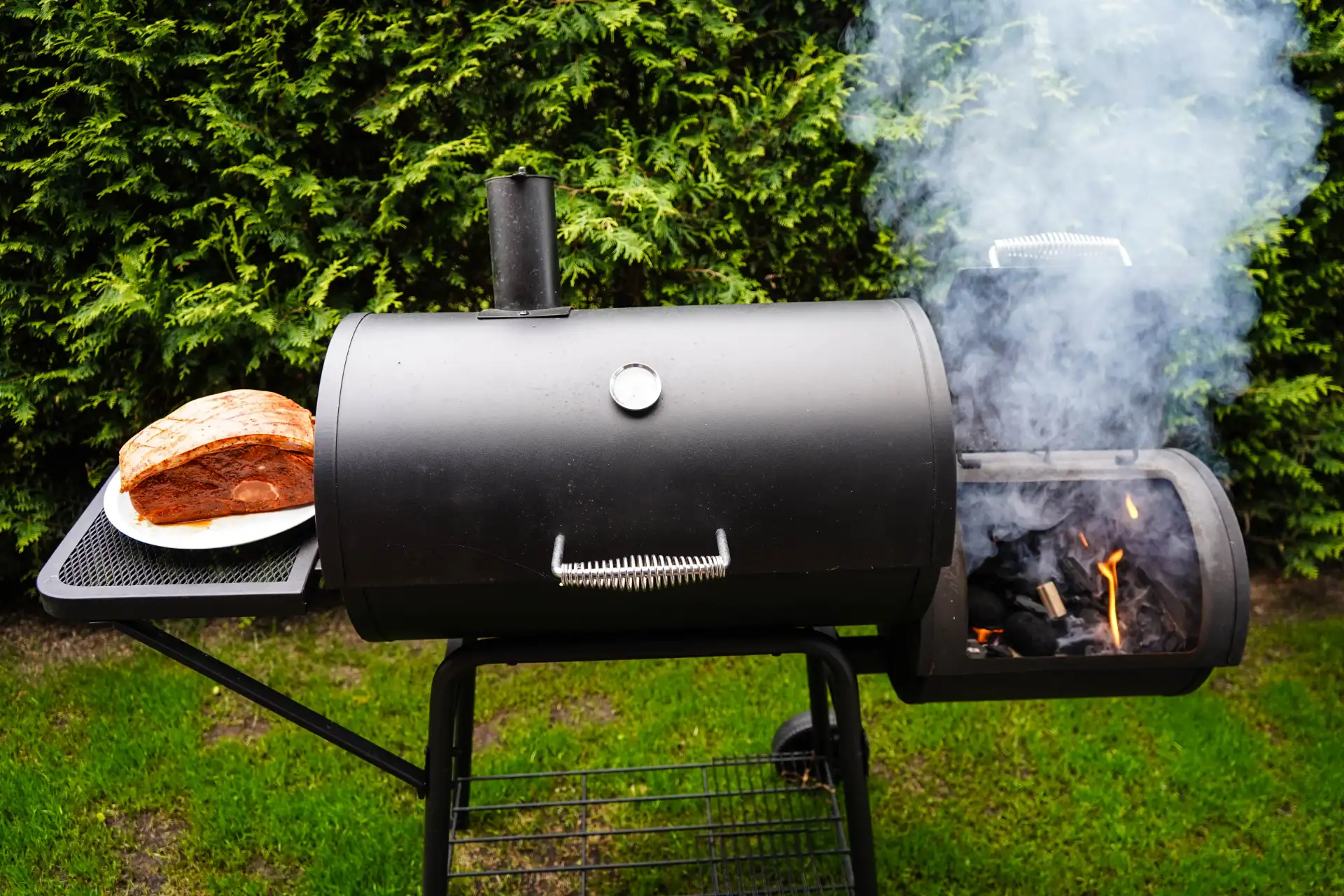 Schwarzer Barrel-Smoker im Garten mit brennendem Holz in der seitlichen Feuerkammer, starkem Rauch und einem Stück Fleisch auf der Ablage